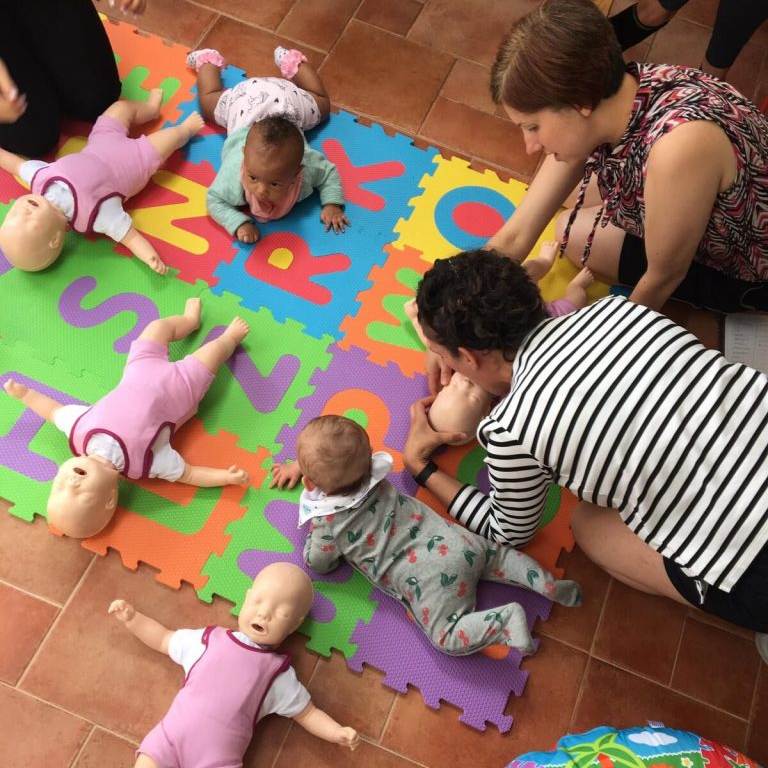Parents perform CPR on resuscitation dummy with their babies safely beside them and a first aid trainer at their side